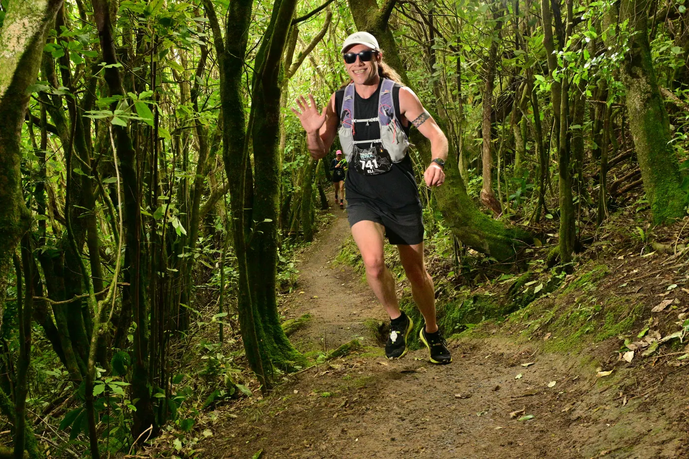 Eli smiling and waving to the camera while running a forest trail race